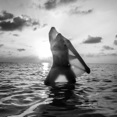 Black White Aufnahme einer Frau. Sie steht im Wasser. Hintergrund ist ein Sonnenuntergang. Man sieht von ihr nichts, weil sie einen transparenten Stoff vor ihren Körper hält.