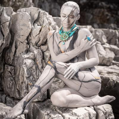 Eine Frau mit geschlossenen Augen sitzt auf einem Felsen und ist so bemalt, dass sie mit dem Hintergrund verschmilzt. Sie trägt eine türkise auffällige Kette mit einer Blume und einen türkisen großen Ring.