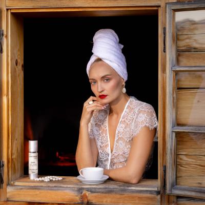 Weiss gekleidete Frau schaut bei Fenster raus mit Tasse und Creme am Fensterbrett