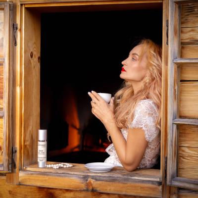 Blonde FRau blickt verträumt mit Tasse in der Hand bei einem Holzfenster hinaus. Davor steht ein Kosmetikprodukt.