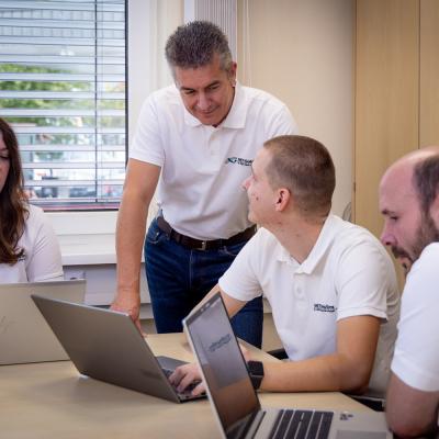 drei Männer und eine Frau unterhalten sich in einem Büro vor dem Laptop