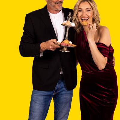 Mann und blonde Frau stehen vor gelbem Hintergrund un nehmen sich Kekse von einem Tablett.