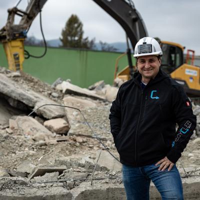 Mann mit weißem Bauhelm am Kopf steht vor schwerem Baugerät und schaut in die Kamera