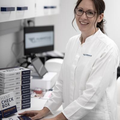 Eine dunkelhaarige Frau mit Brille und weißem Arztmantel steht in einem Labor und lächelt in die Kamera. Sie hat ein Checkbox für Hanf in der Hand.