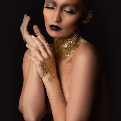 Nacktshooting ohne Details. Frau mit schwarzen Lippen und Goldmakeup hält Hande vor ihr Gesicht und ihren Oberkörper