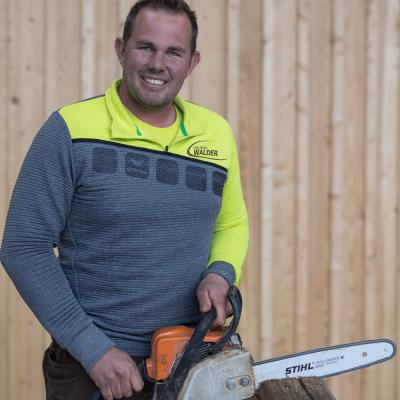 Mann in gelb-grauer Jacke mit Motorsäge in den Händen  lächelt in die Kamera