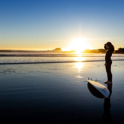 Sonnenuntergang am Meer im Hintergrund. Es herrscht Ebbe und eine Frau steht mit einem Surfbrett daneben und schaut Richtung Sonne