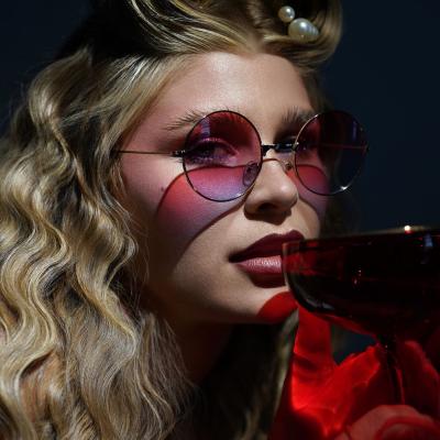 Frau im Retro-Styling mit runder Sonnenbrille hält rotes Glas im dramatischen Licht